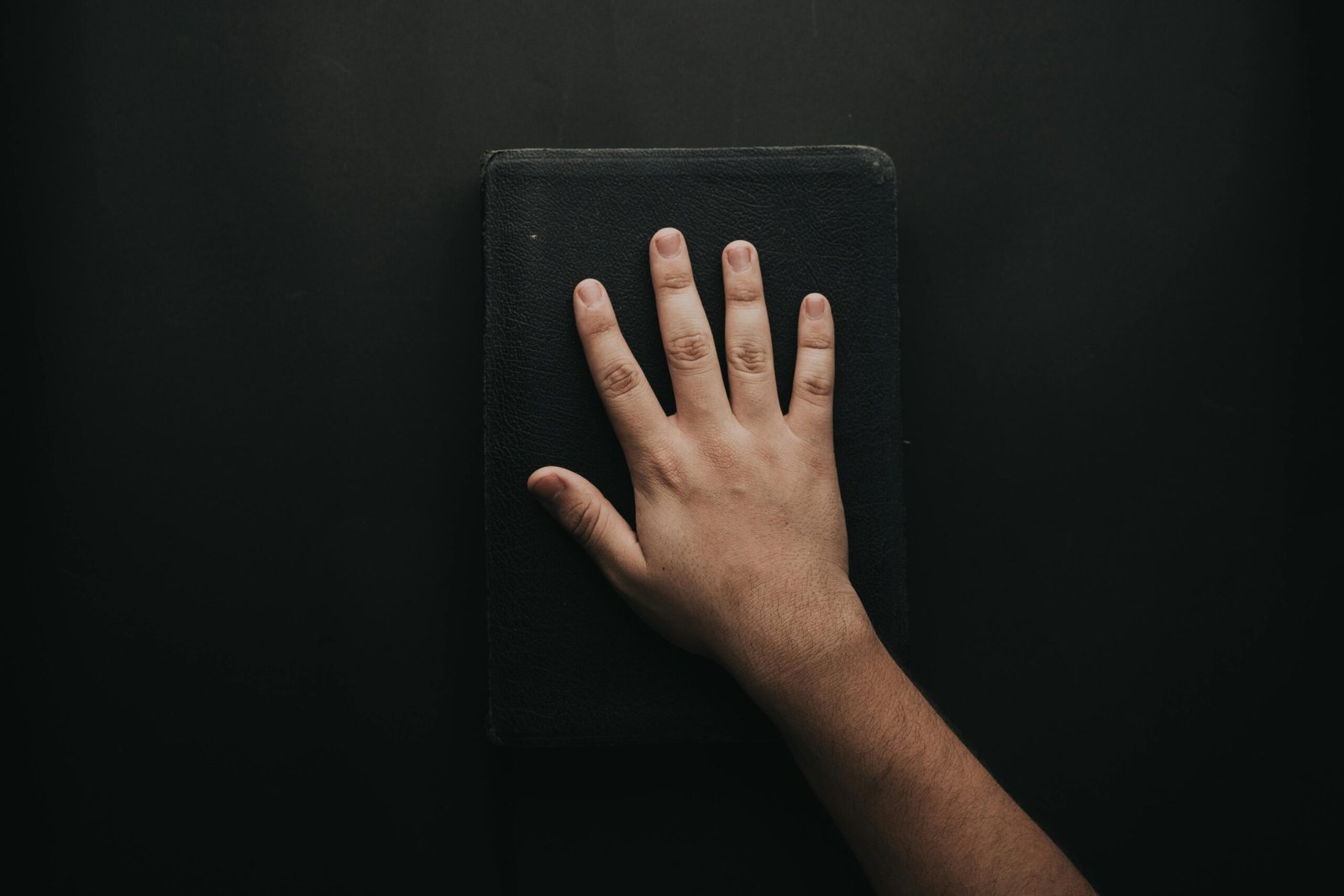 A close-up of a hand resting on a black book against a dark backdrop.